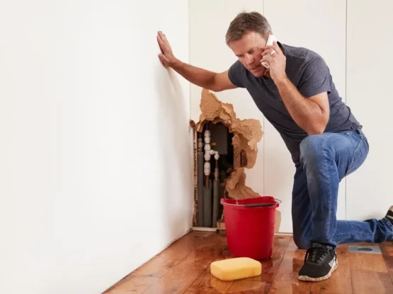 Man kneeling near a damaged wall with exposed plumbing pipes, holding a phone, looking concerned, with a red bucket and sponge nearby, illustrating the challenges of DIY plumbing.