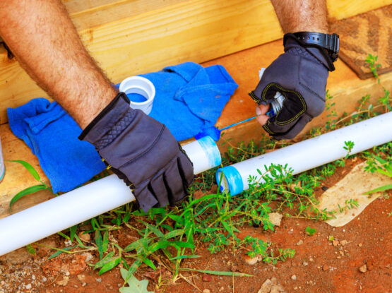Hands wearing gloves working on connecting PVC pipes for water line installation, with tools and blue cloth nearby, in a construction setting.